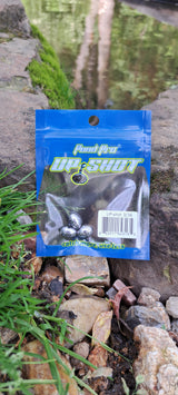 A pack of Up-Shot precision fishing weights placed on a rock near a body of water, with the product visible through a clear plastic section of the packaging.