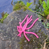 Pond Pro Sqworms in Bubblegum Pink on a rock with moss surrounded by foliage with water in the background.