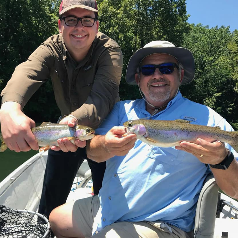 Two men holding rainbow trout on a boat with trees in the background