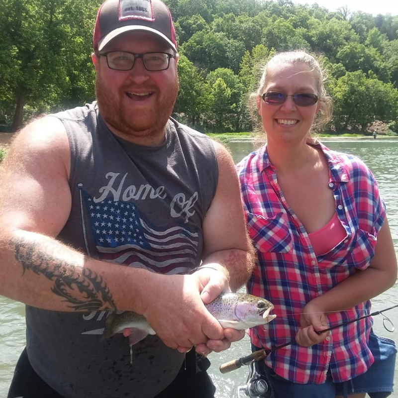 Two people standing by a body of water with trees in the background, one holding a fish.