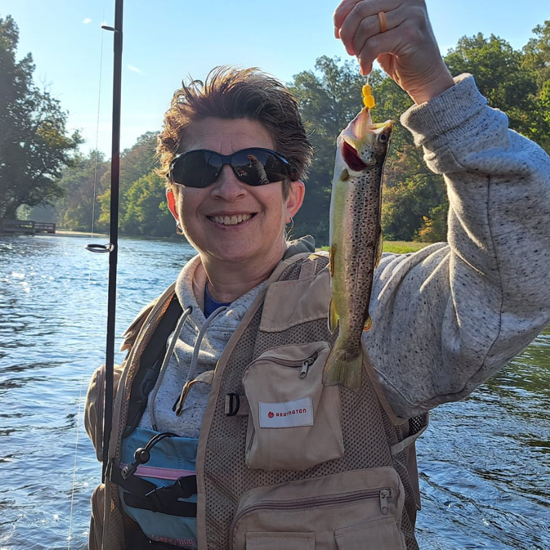 Person holding a fish caught while fishing by a body of water with trees in the background.